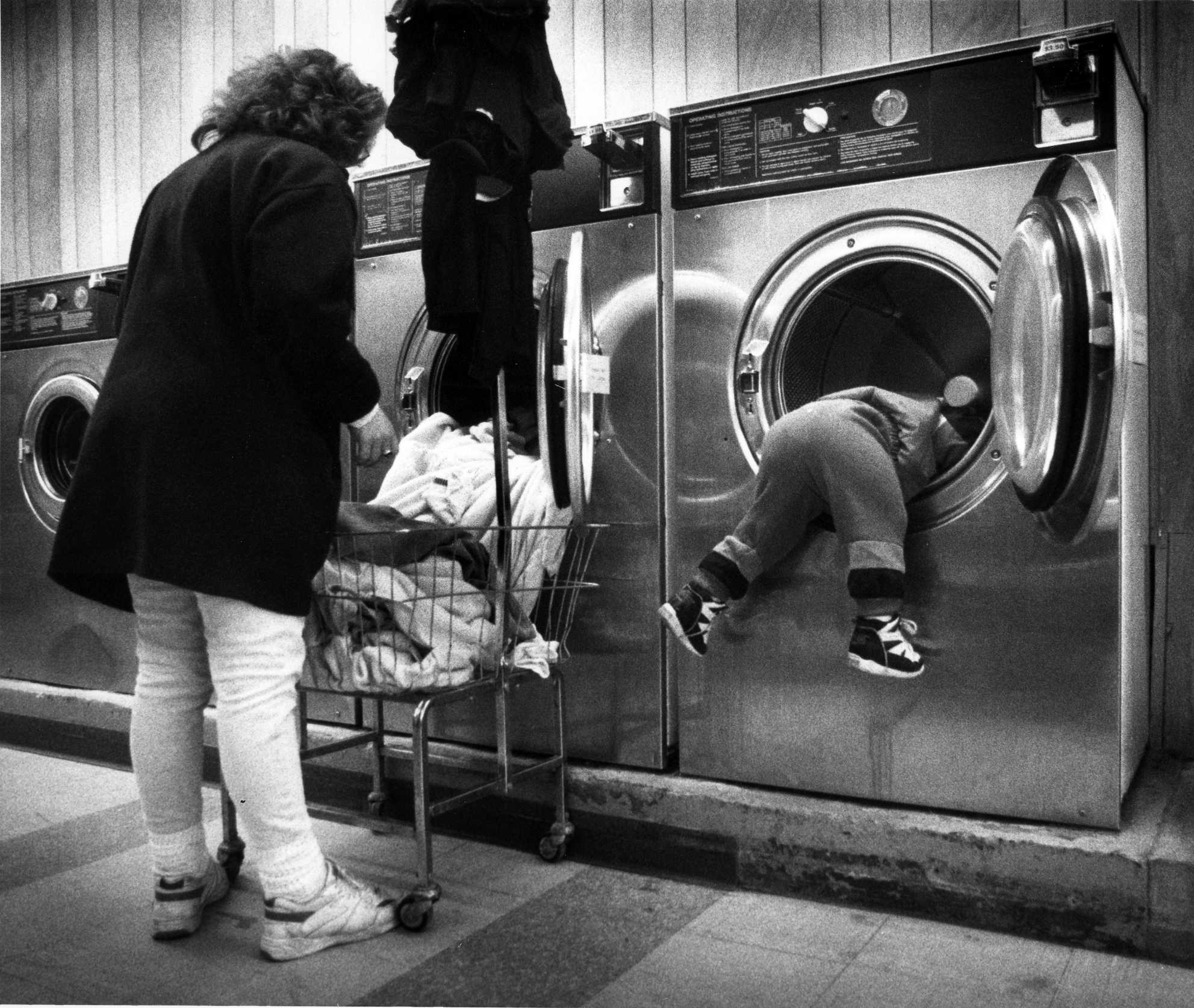 A curious toddler plays in a washing machine as his mother tends to her family's wash at a  local laundromat. 1994