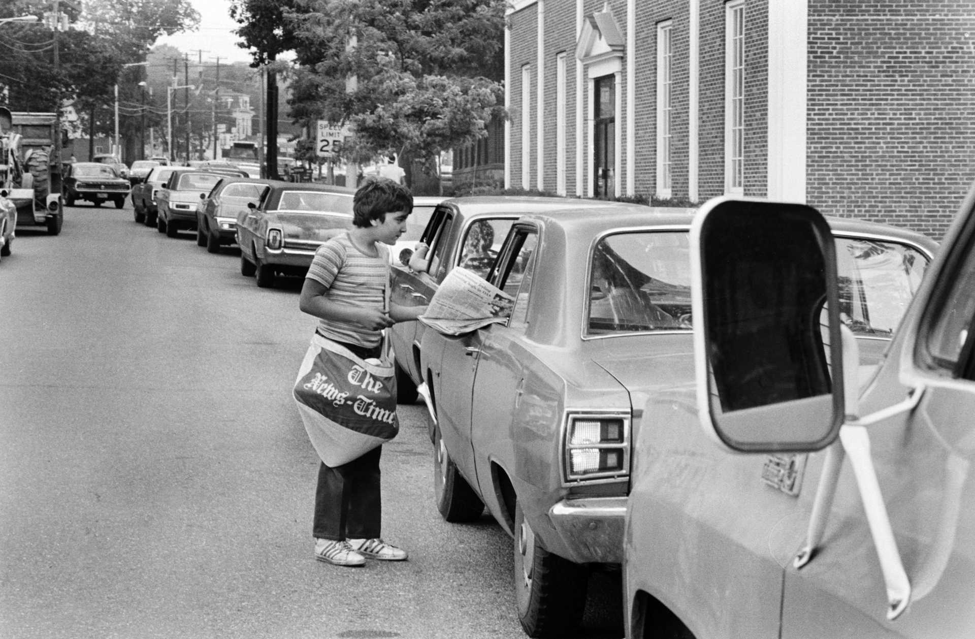 Paperboy Jose Furtado, 12, sells newspapers to people waiting in a line that stretches over a mile, to buy gas on Main Street in Danbury during a summer of national gas shortages. 1979