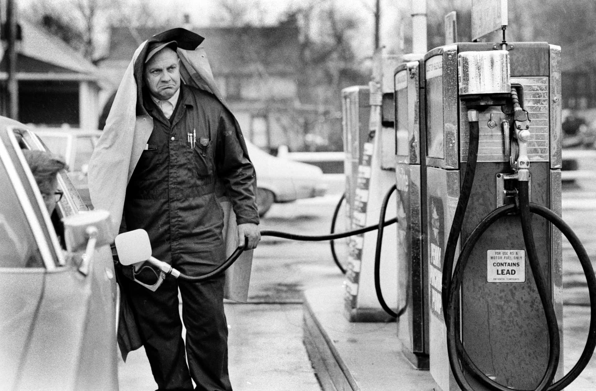 A grumpy gas station attendant protects himself from the rain. 1979.